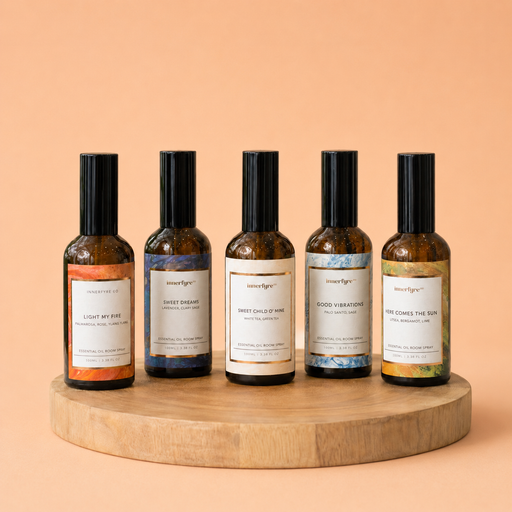 Five amber glass essential oil bottles with black caps and white labels are arranged on a round wooden tray against a warm peach background. The labels read Smokey Eyes, Sweet Dream, Sweet Candy of Mine, Good Vibrations, and You Come the Sun.